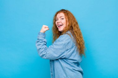 Genç kızıl saçlı kadın kendini mutlu, tatmin olmuş ve güçlü hissediyor, kaslı ve formda, spor salonundan sonra mavi duvara karşı güçlü görünüyor.