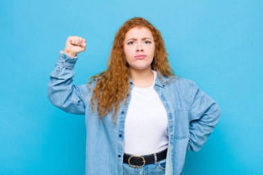 Genç kızıl saçlı kadın ciddi, güçlü ve isyankar hissediyor, yumruğunu kaldırıyor, protesto ediyor ya da mavi duvara karşı devrim için savaşıyor.