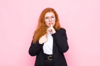 Genç kızıl saçlı kadın ciddi görünüyor ve parmaklarını dudaklarına bastırıp sessizlik ya da sessizlik talep ediyor, pembe duvara karşı bir sır saklıyor.