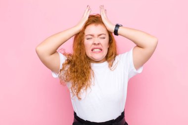 Genç kızıl saçlı kadın stresli ve endişeli, depresif ve hayal kırıklığına uğramış hissediyor, iki elini de pembe duvara doğru kaldırıyor.