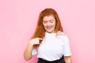 Genç kızıl saçlı kadın neşeyle gülümsüyor, yere bakıyor ve göğsünü pembe duvara gösteriyor.