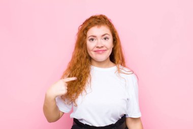 Genç kızıl saçlı kadın gururlu, kendinden emin ve mutlu görünüyor, gülümsüyor ve kendini işaret ediyor ya da pembe duvara karşı bir numara işareti yapıyor.