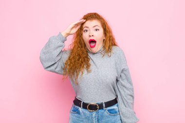 Genç kızıl saçlı kadın mutlu, şaşkın ve şaşırmış görünüyor, gülümsüyor ve pembe duvara karşı inanılmaz ve inanılmaz iyi haberler veriyor.