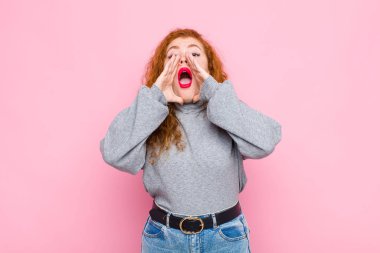 Genç kızıl saçlı kadın mutlu, heyecanlı ve pozitif hissediyor. Elleri ağızda, pembe duvara karşı bağırıyor.