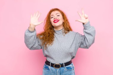 Genç kızıl saçlı kadın gülümsüyor ve arkadaş canlısı görünüyor. Sekiz ya da sekiz numarayı gösteriyor. Elleri önde, pembe duvara doğru sayıyor.