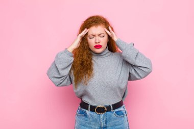 Genç kızıl saçlı kadın stresli ve hayal kırıklığına uğramış görünüyor. Baskı altında çalışıyor. Baş ağrısı çekiyor ve pembe duvara karşı sorunları var.