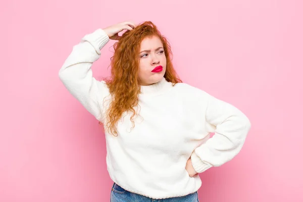 young red head woman feeling puzzled and confused, scratching head and looking to the side against pink wall