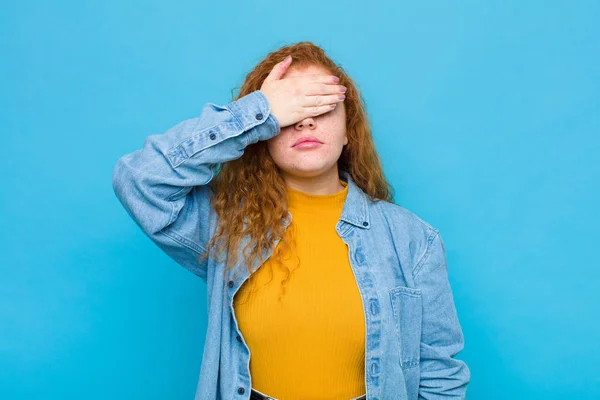 young red head woman covering eyes with one hand feeling scared or anxious, wondering or blindly waiting for a surprise against blue wall