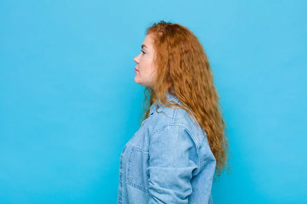 young red head woman on profile view looking to copy space ahead, thinking, imagining or daydreaming against blue wall