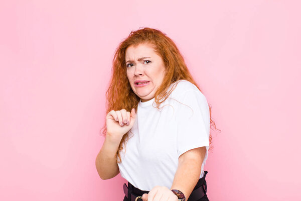young red head woman feeling disgusted and nauseous, backing away from something nasty, smelly or stinky, saying yuck against pink wall