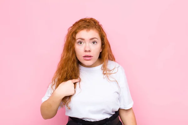 young red head woman feeling confused, puzzled and insecure, pointing to self wondering and asking who, me? against pink wall