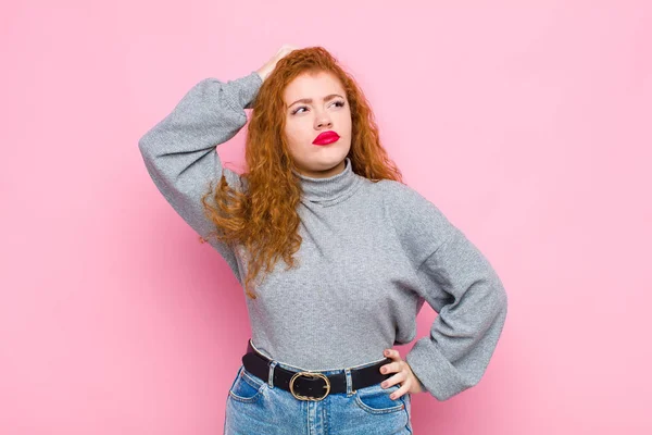 young red head woman feeling puzzled and confused, scratching head and looking to the side against pink wall