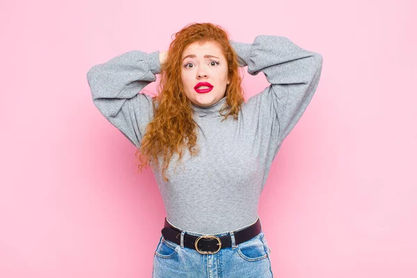 young red head woman feeling stressed, worried, anxious or scared, with hands on head, panicking at mistake against pink wall