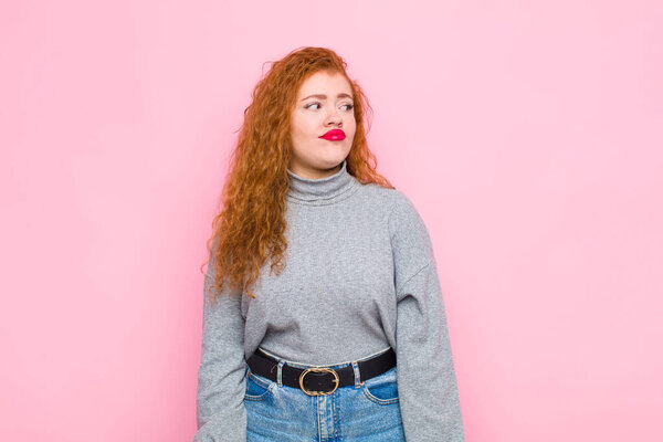 young red head woman wondering, thinking happy thoughts and ideas, daydreaming, looking to copy space on side against pink wall