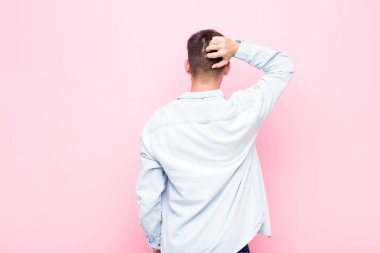 Genç, yakışıklı bir adam bilgisiz ve kafası karışık, bir çözüm düşünüyor, eli kalçasında, diğeri kafasında, pembe duvara karşı dikiz manzaralı.