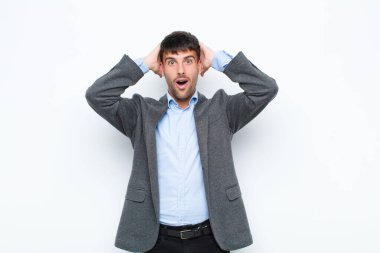 Genç yakışıklı adam heyecanlı ve şaşırmış görünüyor, ağzı açık, iki eli başının üstünde, beyaz duvara karşı şanslı bir kazanan gibi hissediyor.