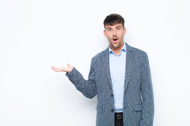 Genç yakışıklı adam şaşırmış ve şok olmuş görünüyor, çenesi düşmüş beyaz duvara karşı açık bir el ile bir nesne tutuyor
