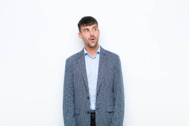 Genç yakışıklı adam şok olmuş, mutlu, şaşırmış ve şaşırmış hissediyor, beyaz duvara karşı ağzı açık bakıyor.