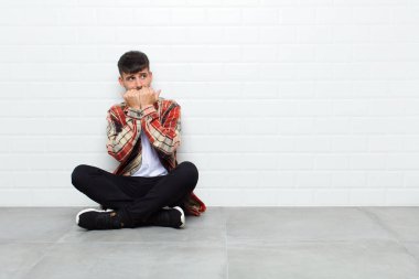 Genç yakışıklı adam endişeli, gergin, stresli ve korkmuş görünüyor, tırnaklarını yiyor ve çimento zeminde oturan yanal fotokopi odasına bakıyor.