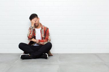 Genç ve yakışıklı bir adam stresli, utanmış ya da üzgün görünüyor, baş ağrısı var, elleri beton zeminde oturuyor.