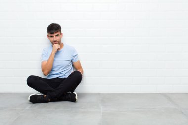 Genç yakışıklı adam ciddi görünüyor, kafası karışık, kararsız ve düşünceli, seçenekler ve seçenekler arasında şüpheci, çimento zeminde oturuyor
