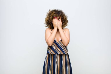 Genç ve güzel bir kadın elleriyle yüzünü kapatıyor. Şaşırmış bir ifadeyle parmaklarının arasından bakıyor ve beyaz duvara bakıyor.