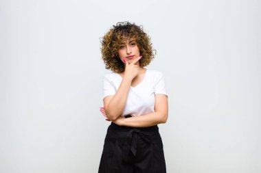 Genç, güzel bir kadın ciddi görünüyor, kafası karışık, kararsız ve düşünceli, beyaz duvara karşı seçenekler veya seçimler arasında şüpheci.