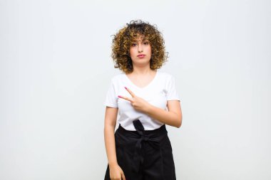 Genç, güzel bir kadın kendini mutlu, pozitif ve başarılı hissediyor. Göğsünde V şeklinde eller yapıyor. Beyaz duvara karşı zafer ya da barış gösteriyor.