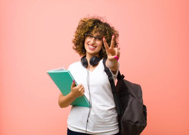 Genç ve güzel bir kız öğrenci gülümsüyor ve mutlu görünüyor, kaygısız ve pozitif, bir eliyle pembe duvara karşı zafer veya barış işareti yapıyor.