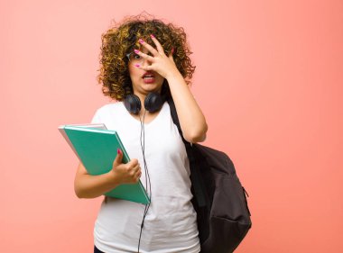 Şok olmuş, korkmuş ya da dehşete düşmüş gibi görünen genç ve güzel bir bayan. Yüzü elle kaplanmış ve parmaklarının arasından pembe duvara bakıyor.