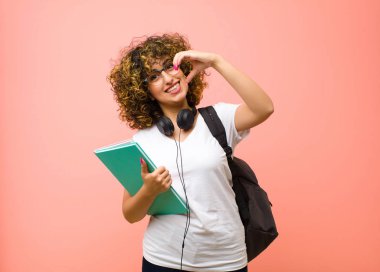 Genç ve güzel bir kız öğrenci gülümsüyor ve mutlu, sevimli, romantik ve aşık, iki elini de pembe duvara dayayıp kalp şekli veriyor.