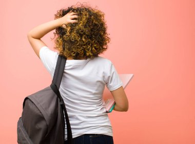 Genç ve güzel bir bayan öğrenci, düşünüyor ya da şüphe ediyor, kafasını kaşıyor, kafası karışık ve şaşkın hissediyor, pembe duvarın arkasında ya da arkasında.