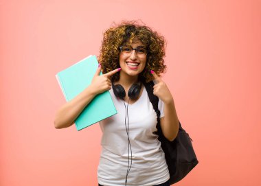 Genç ve güzel bir bayan, kendine güvenerek gülümsüyor. Pembe duvara karşı pozitif, rahat, tatmin olmuş bir tavır sergiliyor.