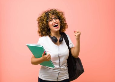 Genç ve güzel bir bayan öğrenci mutlu, şaşırmış ve gururlu hissediyor, başarıyı bağırıyor ve kutluyor pembe duvarda kocaman bir gülümsemeyle.