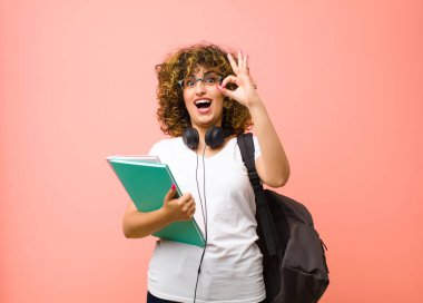 Genç ve güzel bir bayan öğrenci kendini başarılı ve tatmin olmuş hissediyor, ağzı açık gülümsüyor, pembe duvara karşı elle işaret veriyor.