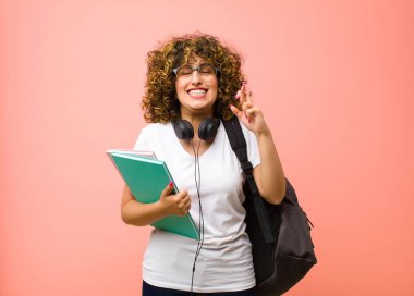 Genç ve güzel bir kız öğrenci. Gülümsüyor ve endişeyle iki parmağını çapraz yapıyor. Endişeli ve pembe duvara karşı iyi şanslar diliyor.