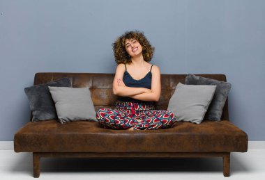 Genç ve güzel bir kadın kollarını kavuşturup mutlu bir şekilde gülüyor, rahat, pozitif ve tatmin olmuş bir pozla koltukta oturuyor..