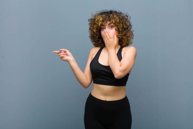 Genç sporcu kadın kendini mutlu, şaşkın ve şaşırmış hissediyor. Ağzını eliyle kapatıyor ve gri duvara karşı yanal fotokopi alanını işaret ediyor.