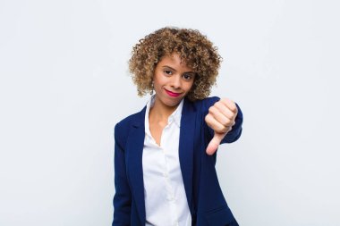 Afro-Amerikan genç kadın, kızgın, sinirli, hayal kırıklığına uğramış ya da hoşnutsuz, düz duvara karşı ciddi bir bakışla başparmaklarını kaldırıyor.