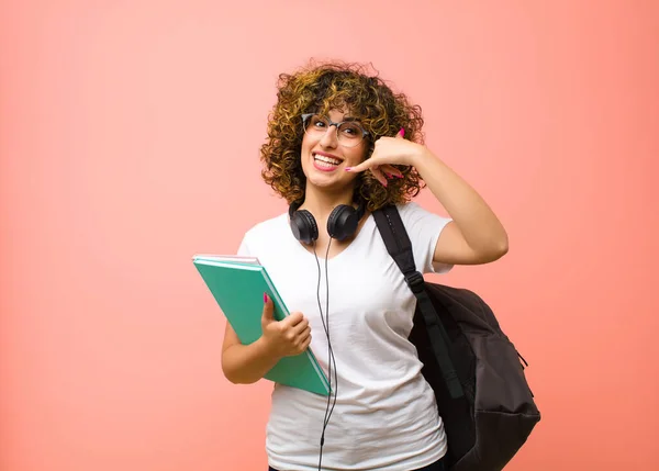 若い学生の女性は笑顔でカメラを指差しながら後でジェスチャーをしてピンクの壁に電話で話します — ストック写真