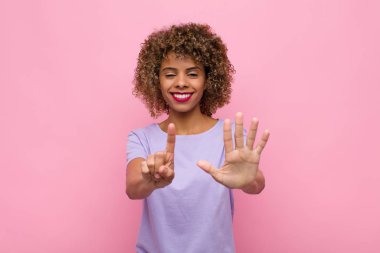 Genç Afrikalı Amerikalı kadın gülümsüyor ve arkadaş canlısı görünüyor. Altı numarayı gösteriyor. Elleri önde, pembe duvara doğru sayıyor.