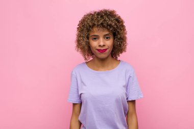 Genç Afrikalı Amerikalı kadın pozitif ve kendinden emin gülümsüyor. Tatmin olmuş, arkadaş canlısı ve pembe duvara karşı mutlu görünüyor.