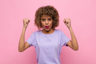 Genç Afrikalı Amerikalı kadın inanılmaz bir başarıyı kutluyor. Kazanan gibi. Heyecanlı ve mutlu görünüyor. Pembe duvara yaslan