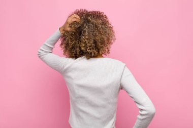 Genç Afrikalı Amerikalı kadın kendini cahil ve kafası karışık hissediyor, bir çözüm düşünüyor, eli kalçasında ve diğerinin kafasında, pembe duvara karşı dikiz penceresi