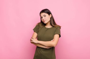 Genç, güzel bir kadın omuz silkiyor, kafası karışık ve kararsız, kolları bağlı ve şaşkın pembe duvara bakıyor.