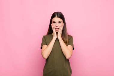 Genç ve güzel bir kadın şok olmuş ve korkmuş hissediyor. Ağzı açık ve elleri pembe duvarda.