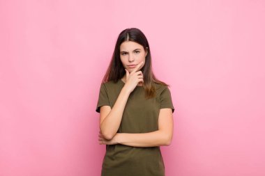 Genç ve güzel bir kadın ciddi, düşünceli ve güvensiz görünüyor, bir kolu çapraz ve çene üzerinde, pembe duvarla seçenekleri tartıyor