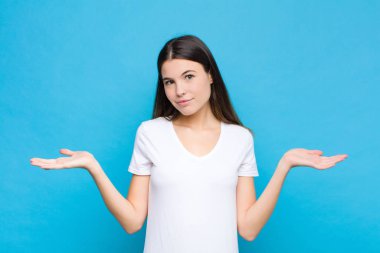 Genç ve güzel bir kadın şaşkın ve şaşkın hissediyor, şüpheci, farklı seçenekleri tartıyor ya da seçiyor mavi duvara karşı komik bir ifadeyle