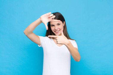 Genç, güzel bir kadın mutlu, arkadaş canlısı ve pozitif hissediyor, gülümsüyor ve elleri mavi duvara dayalı bir portre ya da fotoğraf çerçevesi yapıyor.
