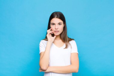 Genç ve güzel bir kadın şok ve inançsızlık içinde, elleri yanağında ve kolları çaprazlanmış, sersemlemiş ve mavi duvara karşı şaşkınlık içinde.